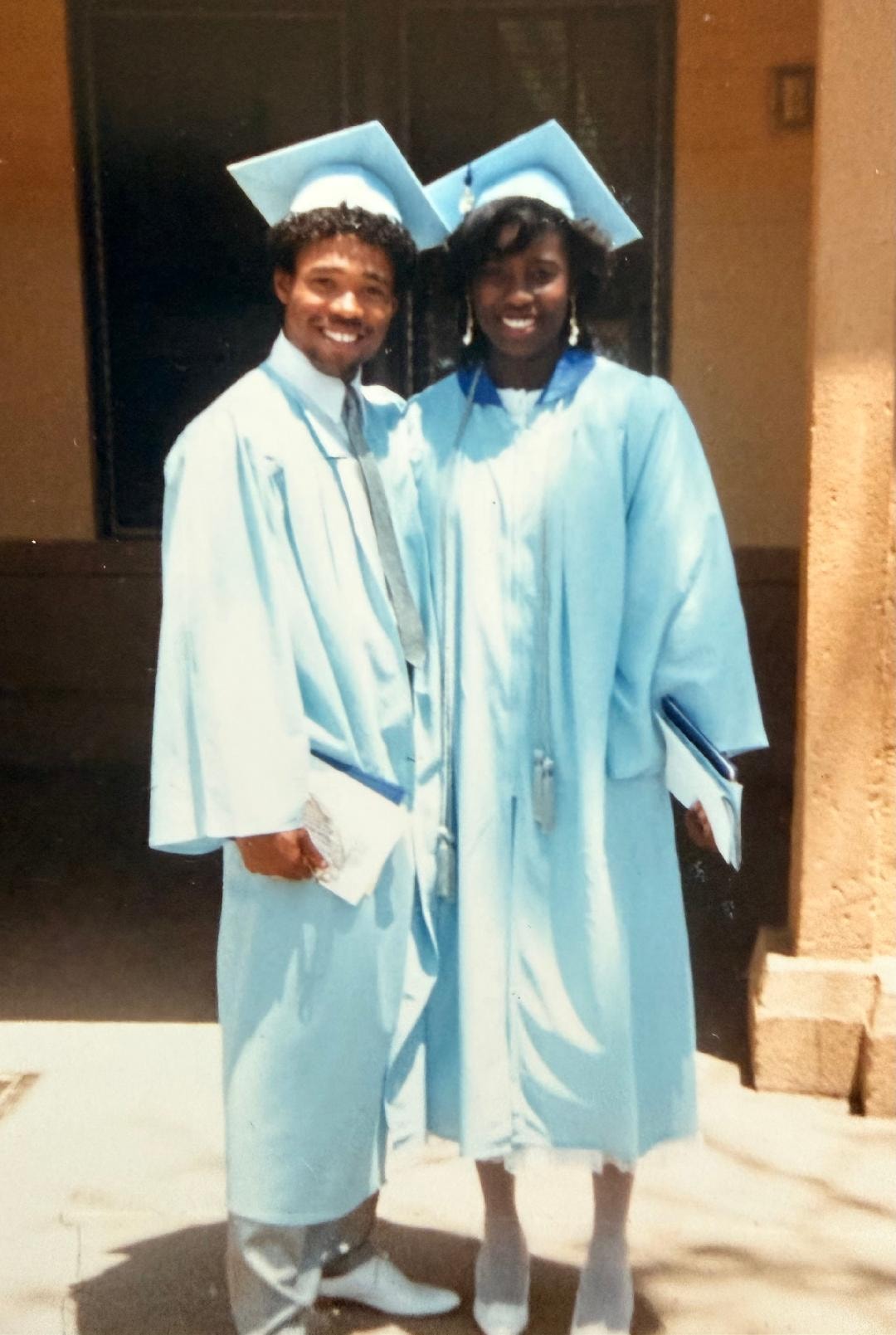 Two graduates in light blue caps and gowns posing together on graduation day