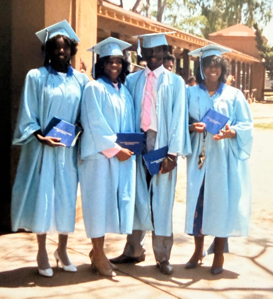 Four graduates in light blue caps and gowns holding diplomas on graduation day